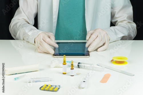 Modern MD office table reception. Medical doctor in gloves and lab coat at white office table with medical utensils using tablet PC to manage consultation timetable photo