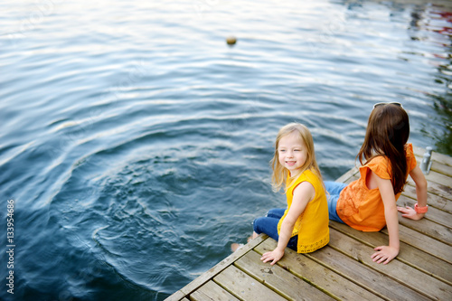 Two cute little girls sitting on a wooden platform by the river or lake