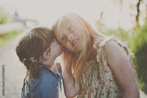 Side view of girl whispering into sister's ear on footpath photo