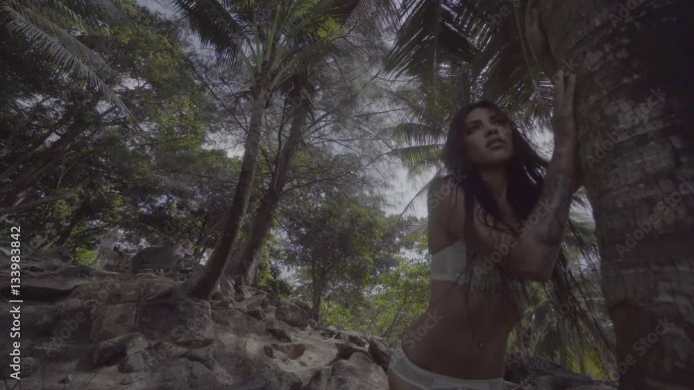 Down view of sensual brunette beauty wearing mint underwear with wet hair, looking like amazon woman, walking close to the tree and looking somewhere over tropical green jungle and sky background Down view of sensual brunette beauty wearing mint underwear with wet hair, looking like amazon woman, walking close to the tree and looking somewhere over tropical green jungle and sky background