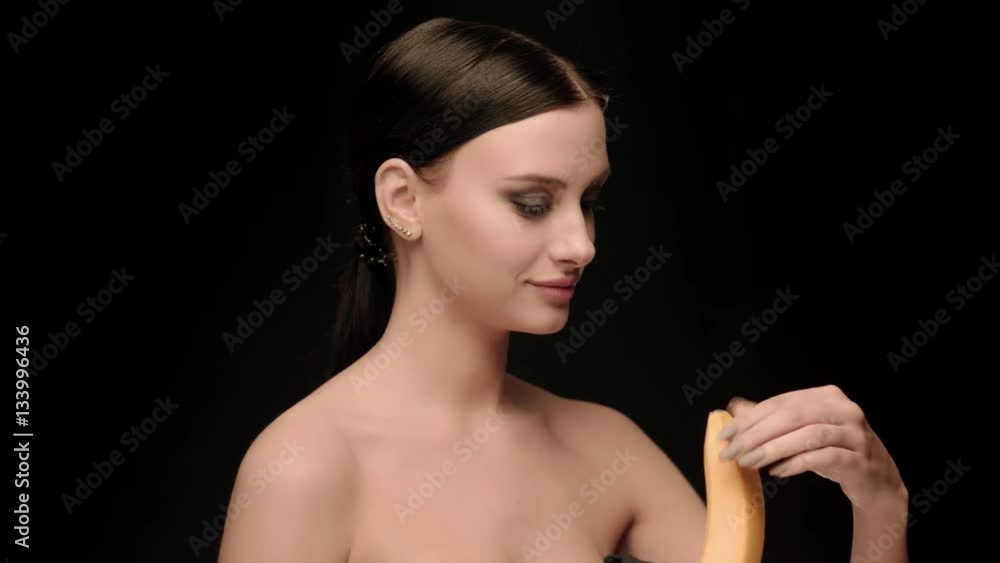 Fashion brunette standing on black wall background in studio, eating a banana Fashion brunette standing on black wall background in studio, eating a banana