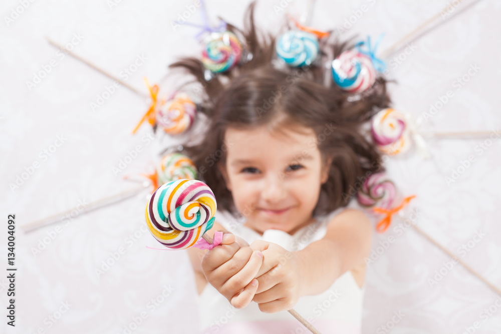 Pretty girl among the sweets, candies and lollipops. Funny child with candy lollipop, happy little girl eating big sugar lollipop, kid eat sweets. surprised child with candy. 