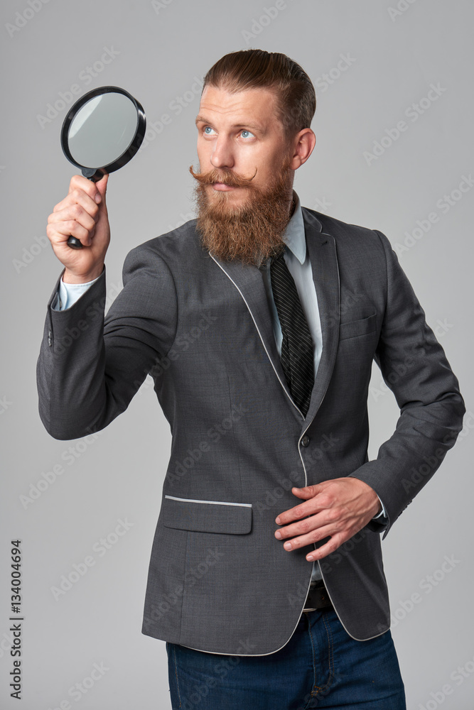 Serious hipster business man with beard and mustashes in suit looking up through magnifying glass, over grey background Serious hipster business man with beard and mustashes in suit looking up through magnifying glass, over grey background
