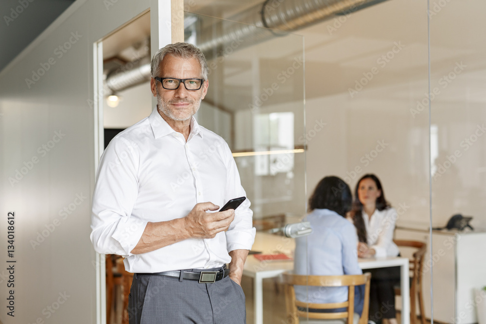 Confident businessman with cell phone in office with employees in background Confident businessman with cell phone in office with employees in background