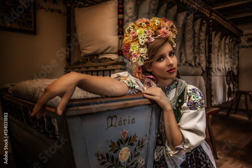 Woman dressed in slovak folk dress sitting indoor in the antique bedroom leaning towards a traditional old style crib photo