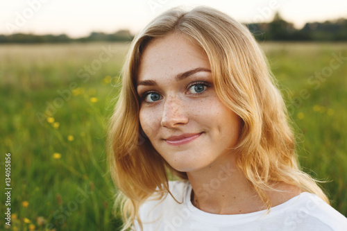 A portrait of beautiful young blue-eyed girl with light hair having charming smile and dimple on her face looking into camera standing over green background having nice time relaxing outdoor in summer photo