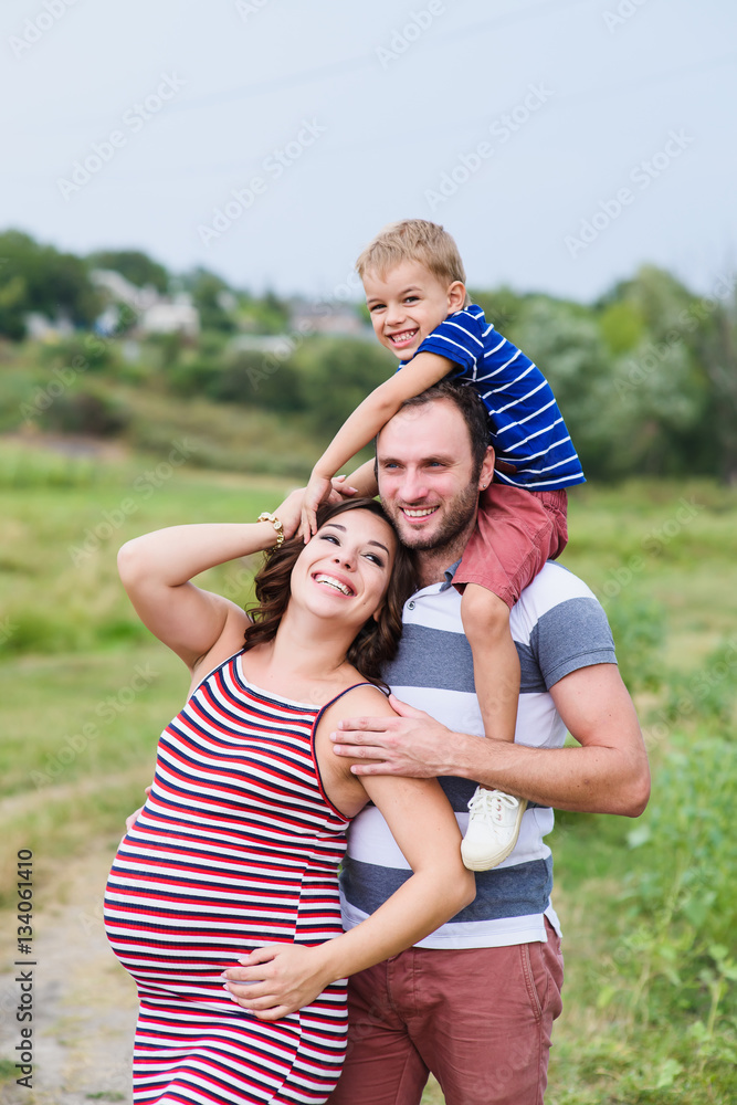Pregnant family of three in expectation of baby. Father mother and son playing together in day outside. Happy people hugging. Smiling laughing baby sitting on shoulders of daddy. Pregnant family of three in expectation of baby. Father mother and son playing together in day outside. Happy people hugging. Smiling laughing baby sitting on shoulders of daddy.