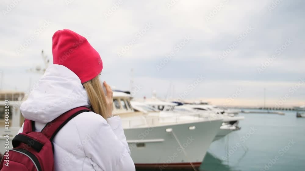 Young beautiful caucasian girl standing on a pier with boats and talks to the mobile phone Young beautiful caucasian girl standing on a pier with boats and talks to the mobile phone