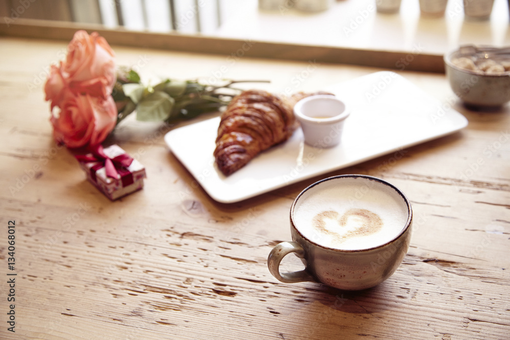 Romantic breakfast, Valentine's Day celebrating. Present box, rose flowers, fresh croissant, coffee on wooden table. Focus on cup. Daylight from window. Romantic breakfast, Valentine's Day celebrating. Present box, rose flowers, fresh croissant, coffee on wooden table. Focus on cup. Daylight from window.