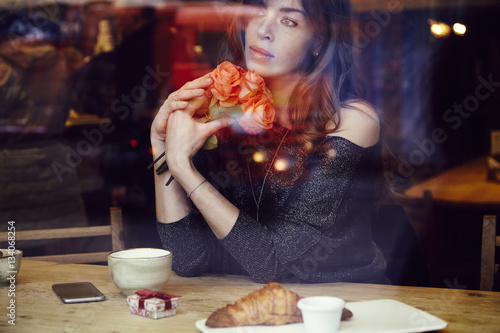 Beautiful woman with long hair holding red roses flowers in hand look through the window in cafe. Coffee and croissant on wooden table. St. Valentine s Day or Birthday concept.