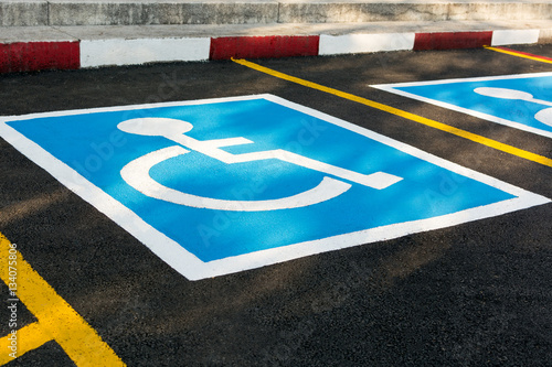 Close up of empty handicapped reserved parking space with wheelchair symbol on black asphalt. No parking white painted letters and blue diagonal lines in background. Rough cracked pavement.