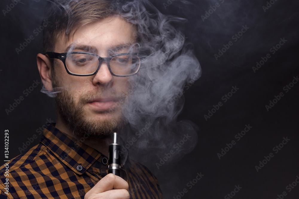man with a beard wearing glasses on black background smoke Vape. large portrait of . electronic cigarette. E- for vaping. Popular devices the year - modern device. Stop smoking, man with a beard wearing glasses on black background smoke Vape. large portrait of . electronic cigarette. E- for vaping. Popular devices the year - modern device. Stop smoking,