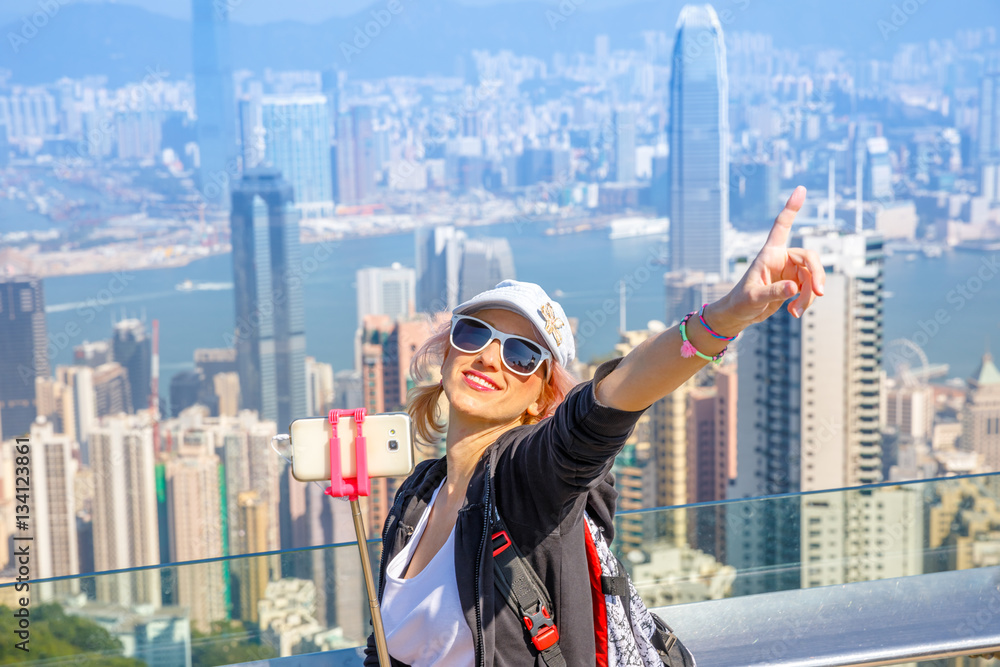 Naklejka premium Koncepcja azji wolności podróżnika. Hongkong Victoria Peak turysta robi zdjęcie selfie stick ze smartfonem z widokiem na Port Wiktorii z Peak Tower. Niewyraźne tło.