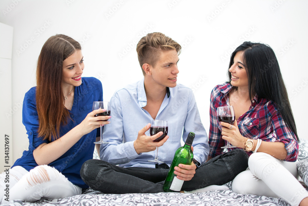 Three young friends sitting on bed and drinking red wine together.