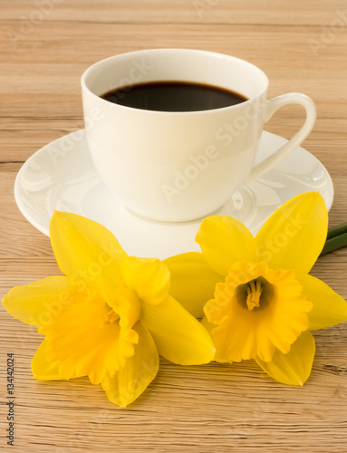 A cup of coffee with two daffodils in the foreground on a brown wooden background.  