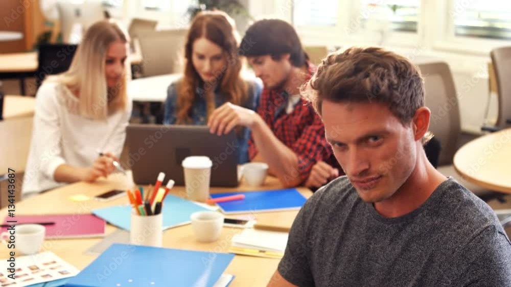 Male business executive smiling at camera while colleagues interacting at desk Male business executive smiling at camera while colleagues interacting at desk