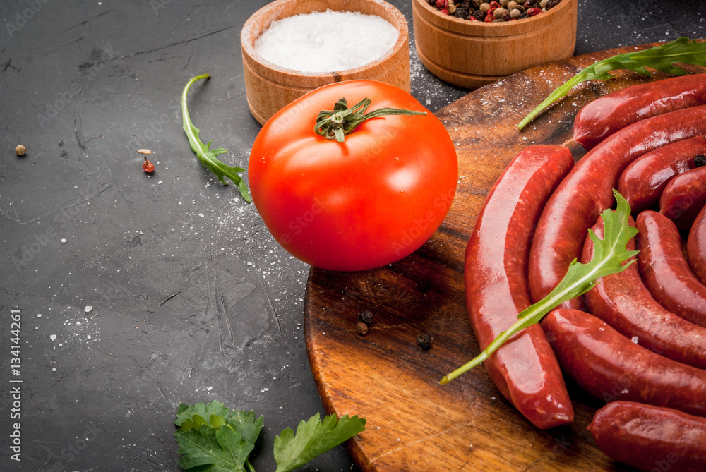Homemade raw beef sausages, near the herbs and spices for cooking vegetables. On a cutting board, on a dark gray stone table, close view, copy space Homemade raw beef sausages, near the herbs and spices for cooking vegetables. On a cutting board, on a dark gray stone table, close view, copy space