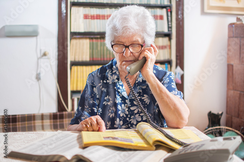 Senior woman talking on phone and searching yellow pages for services.