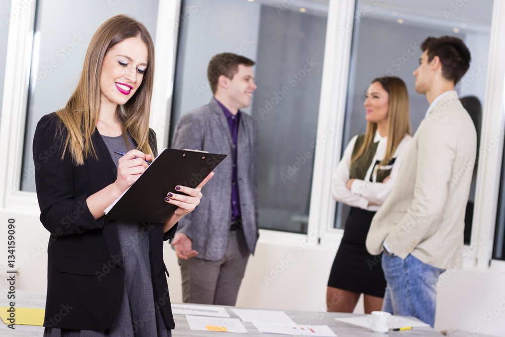Beautiful modern business woman holding a clipboard in the office with colleagues in the background.