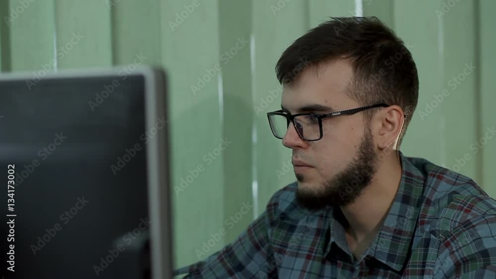 a young man with a beard and glasses works behind a computer a young man with a beard and glasses works behind a computer