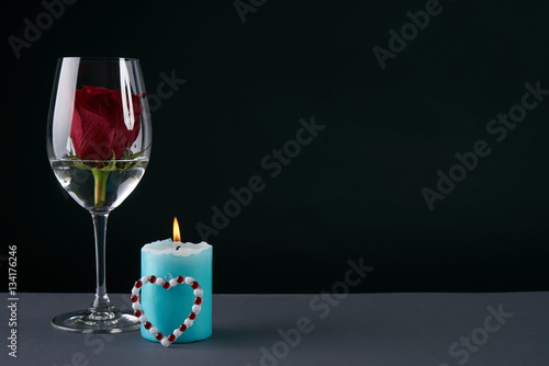 Wineglass with rosebud inside, candle and heart on dark background. Love card concept with copy space. Valentine's day theme
