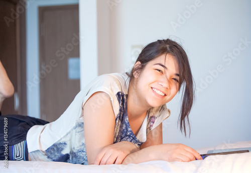 Biracial teen girl lying on bed smiling, using tablet computer