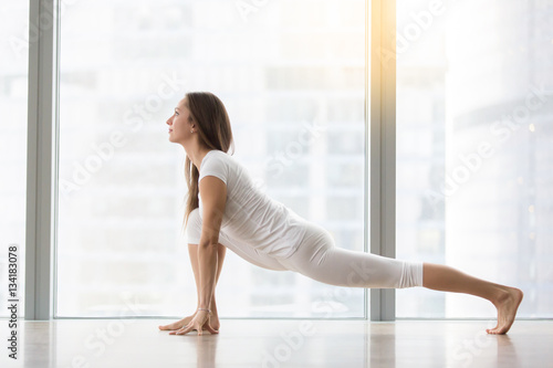 Young attractive happy woman practicing yoga, stretching in Lizard exercise, Utthan Pristhasana pose, working out, wearing sportswear, white t-shirt, pants, full length, at floor window with city view