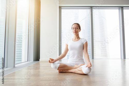 Fototapeta Naklejka Na Ścianę i Meble -  Young attractive woman practicing yoga, sitting in Ardha Padmasana exercise, Half Lotus pose, working out, wearing white t-shirt, pants, meditation session at floor window with city view. Full length