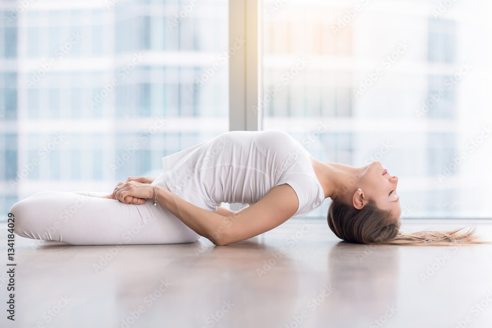 Young attractive woman practicing yoga, stretching in Fish exercise, variation of Matsyasana with lotus legs pose, working out, wearing sportswear, white t-shirt, pants, full length, near floor window Young attractive woman practicing yoga, stretching in Fish exercise, variation of Matsyasana with lotus legs pose, working out, wearing sportswear, white t-shirt, pants, full length, near floor window