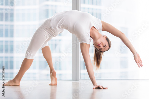 Young attractive woman practicing yoga, stretching in Wild Thing, Flip-the-Dog, exercise, Camatkarasana pose, working out, wearing sportswear, white t-shirt, pants, indoor full length, floor window