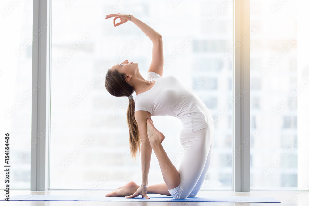 Young happy attractive woman practicing yoga, sitting in Camel exercise, Ustrasana pose, mudra, working out, wearing sportswear, white t-shirt, pants, indoor full length, floor window with city view Young happy attractive woman practicing yoga, sitting in Camel exercise, Ustrasana pose, mudra, working out, wearing sportswear, white t-shirt, pants, indoor full length, floor window with city view