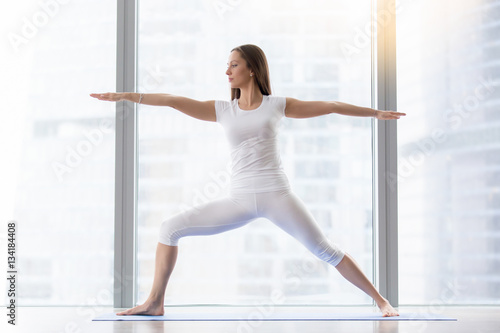 Young attractive woman practicing yoga, standing in Warrior two exercise, Virabhadrasana II pose, working out, wearing sportswear, white t-shirt, pants, indoor full length, floor window with city view