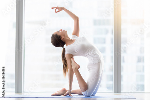 Young happy attractive woman practicing yoga, sitting in Camel exercise, Ustrasana pose, mudra, working out, wearing sportswear, white t-shirt, pants, indoor full length, floor window with city view