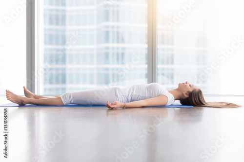 Young woman practicing yoga, lying in Savasana exercise, Dead Body, Corpse pose, working out, wearing sportswear, white t-shirt, pants, indoor full length, near floor window with city view, side view