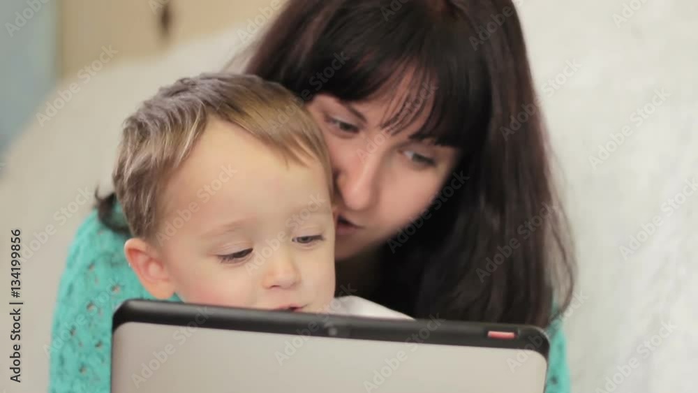 Mother and child looking at the display of the tablet while sitting on a couch at home Mother and child looking at the display of the tablet while sitting on a couch at home