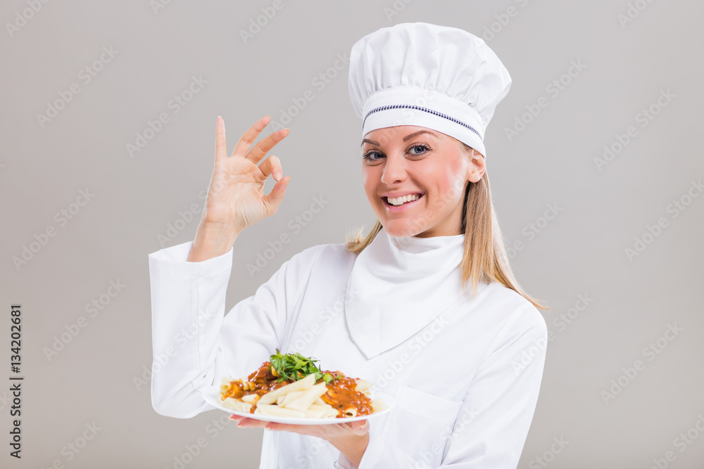 Beautiful female chef is showing ok sign and prepared meal on gray background.
Beautiful female chef is showing ok sign and prepared meal on gray background.