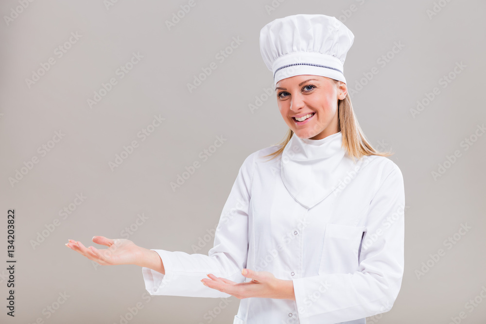 Portrait of beautiful female chef showing welcome gesture on gray background.
Portrait of beautiful female chef showing welcome gesture on gray background.