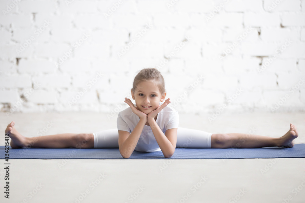 Funny portrait of cute girl child practicing yoga, doing splits, Samakonasana stretching exercise, Straight Angle pose, working out wearing sportswear, indoor full length, white loft studio background