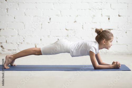 Serious girl child practicing yoga, standing in Dolphin Plank exercise, phalankasana pose working out wearing sportswear, t-shirt, pants, indoor full length, white loft studio background