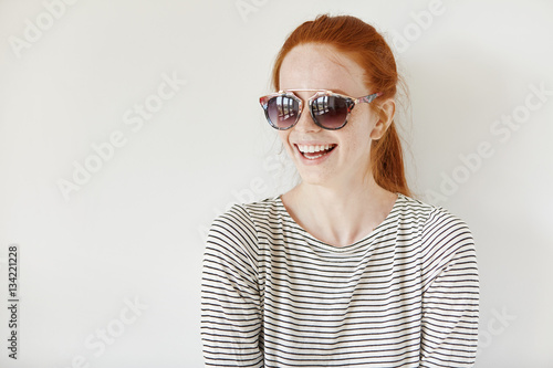 Fototapeta Naklejka Na Ścianę i Meble -  Cheerful hipster girl with freckles and ginger hair wearing stylish shades and sailor shirt smiling happily, having positive mood, standing isolated against white wall with copy space for your content