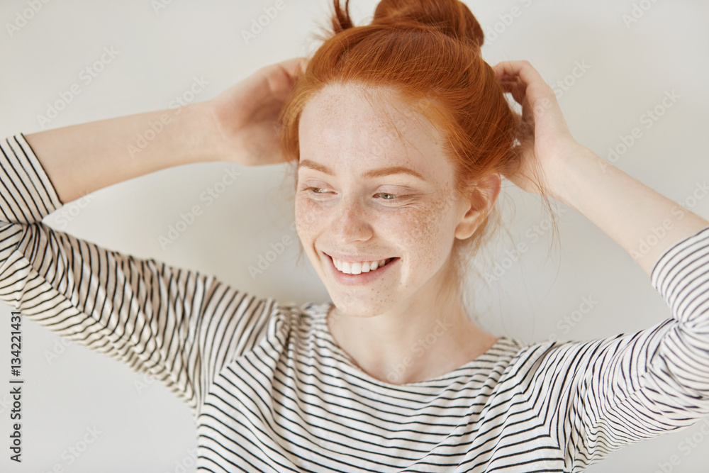 Fototapeta premium People and lifestyle concept. Attractive young woman with ginger hair and freckled skin wearing striped top smiling happily while adjusting her hairstyle before going out to party with her friends