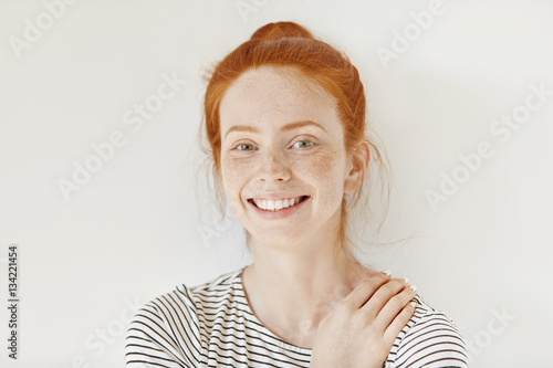 Stylish freckled teenage girl with hair knot having fun indoors and looking at camera with cheerful smile. Beautiful young redhead lady smiling happily keeping hand on her breast, isolated on white