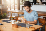 Fashionable young bearded man in black hat checking his newsfeed on social media and listening to songs online on earphones, using music app on his cell phone, enjoying free wi-fi during lunch at cafe