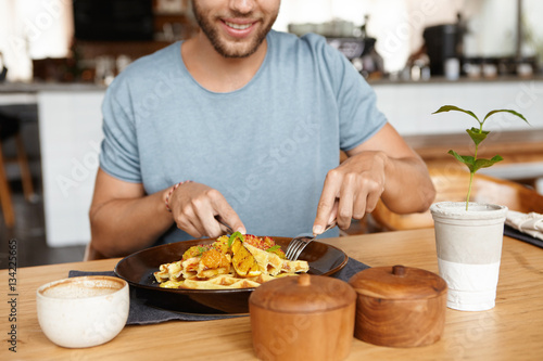 Cropped portrait of happy young bearded male in t-shirt smiling cheerfully while enjoying tasty meal during lunch at cozy restaurant, sitting at wooden table, eating food with knife and fork