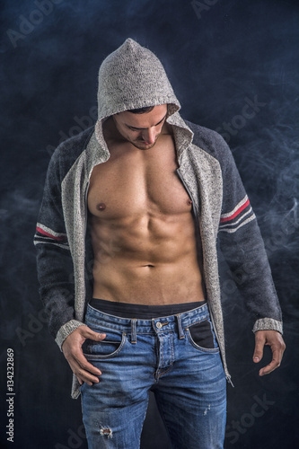 Confident, attractive young man with open vest on muscular torso, ripped abs and pecs. On dark background