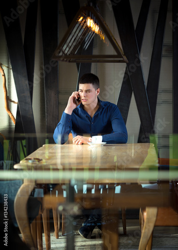 Portrait of a handsome young man on a coffee break in a restaurant. He is talking on the phone and having a cup of coffee.