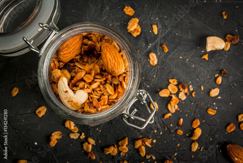 Golden granola cereal flakes with dried fruits, nuts and honey in a glass jar. On a dark stone table. copy space, top view