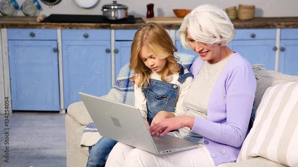 Granny and her little girl using laptop computer at home. Pretty granddaughter helping her granny to log in while sitting on sofa or couch at home. Granny and her little girl using laptop computer at home. Pretty granddaughter helping her granny to log in while sitting on sofa or couch at home.