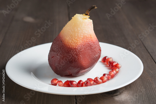 one poached pear on white plate with red pomegranate seeds on dark wooden background