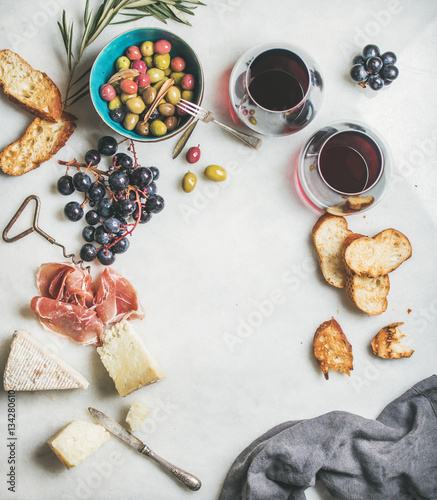 Wine and snack set. Variety of cheese, olives, prosciutto, roasted baguette slices, black grapes and glasses of red wine over grey marble background, top view, copy space